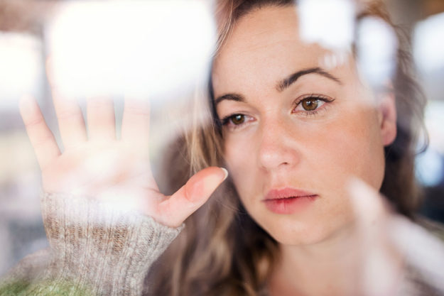 a woman stares pensively out the window with her hand up against glass as she thinks about depression treatment during covid 19