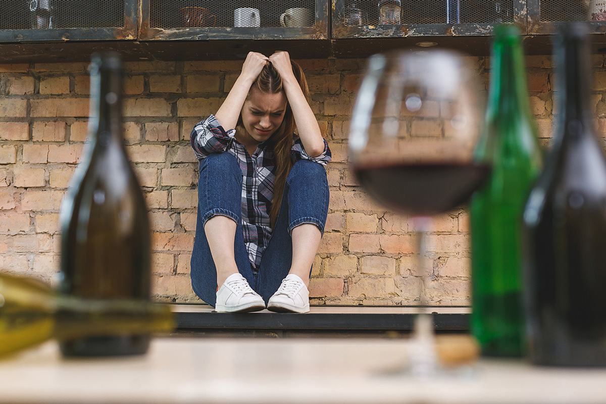 a woman sits beneath a kitchen island as she considers the risks of alcohol abuse